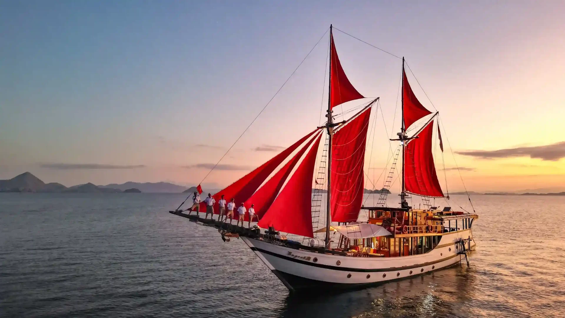Phinisi with red sails and crew on the bowsprit at sunset