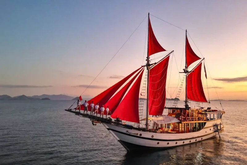 Phinisi with red sails and crew on the bowsprit at sunset